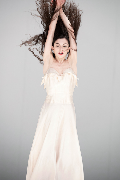 a woman in a slinky white gown with thin straps appears to hang in the air or vertically float. Facing us, her arms are lifted above her head, and her hair flies along with them.